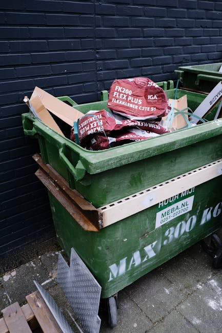 A large green wheelie bin filled with several red and black flexible pipe connectors, positioned against a dark blue brick wall on a paved outdoor surface. The connectors are stacked loosely inside the bin, with some resting on top and partially protruding. The bin is situated on three caster wheels, with a white polymer tray attached below the rim, serving as a protective cover. Nearby, part of another green bin is visible, also placed on the paved ground, indicating an outdoor rubbish collection or waste removal setting. The scene reflects an environment suitable for private waste disposal or an alternative to traditional council bin collections, with the branding 'Rubbish Removal Uxbridge' subtly implied by the context of rubbish management services.