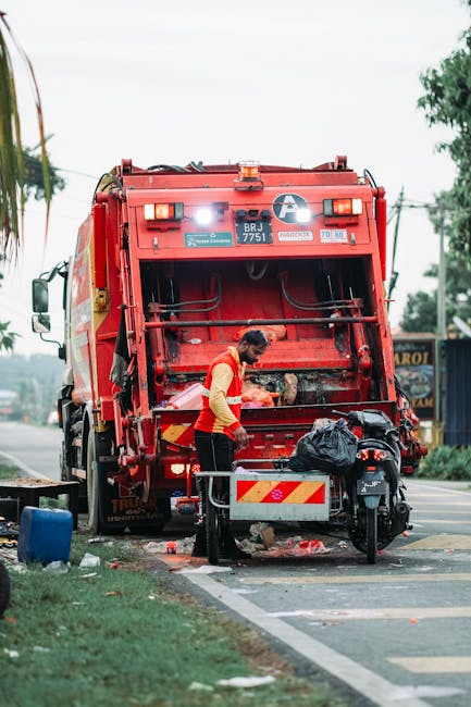 A sanitation worker with a dark ponytail, wearing a high-visibility orange vest over a long-sleeved yellow shirt, is operating a large red rubbish collection vehicle parked on the side of a paved road. The back of the vehicle features mechanical arms and a large open compartment used for collecting waste, with various warning and identification labels visible on its surface. The worker appears to be handling rubbish or inspecting the contents, which are being loaded or processed. In front of the vehicle, a black garbage bag rests on a small metal platform attached to a motorized waste collection tricycle, positioned on the road's edge. Nearby, a blue container and some scattered debris are visible on the pavement. The scene is set outdoors with greenery, trees, and utility poles in the background, under a cloudy sky, indicating a routine waste collection activity associated with private rubbish removal services like Rubbish Removal Uxbridge. The environment suggests a typical urban or suburban street scene where waste collection is occurring, supporting clean-up and rubbish clearance efforts.