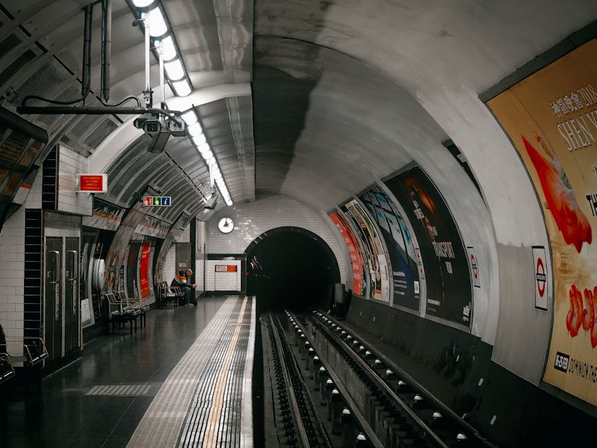 A view of an underground metro station platform with curved white tiled walls and a dark tunnel entrance at the center. The platform features a tactile paving strip along the edge for safety, and a metal bench with a passenger seated on it. On the right side, colourful advertisements with Chinese characters are displayed on the curved wall. Overhead, fluorescent lighting illuminates the station, and a clock showing the time is mounted on the wall. The station is equipped with safety signs, including a red emergency alarm box and directional indicators, and the overall environment appears clean and modern with a minimalist design suitable for urban public transport. This scene provides a clear visual connection to underground transportation systems and the necessity of waste management and cleaning services in public infrastructure facilities such as the one operated by Rubbish Removal Uxbridge, highlighting the importance of maintaining cleanliness even in transit environments.